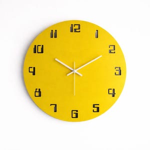 Pode incluir: Um relógio de parede redondo amarelo com algarismos pretos e ponteiros brancos. O mostrador do relógio é amarelo brilhante e os números estão em uma fonte moderna. Os ponteiros finos e brancos indicam as horas. Design minimalista.