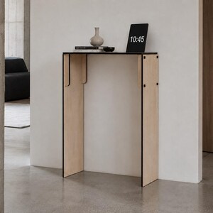 May include: A tall, narrow console table constructed from light-colored plywood panels with exposed black edges. The table features a flat rectangular top and two vertical side supports. A small vase, books, and a digital clock displaying 10:45 sit on top.