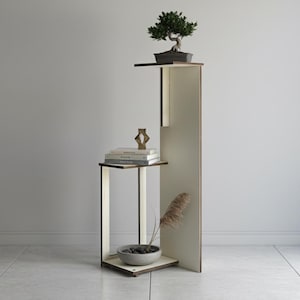 Op de afbeelding: Een moderne plantenstandaard met meerdere niveaus, met een bonsai-boom op de bovenste plank. De standaard heeft een crèmekleurige afwerking met donkere houten accenten. Een kleine kom en boeken staan op de onderste plank, ongeveer 150 cm hoog.