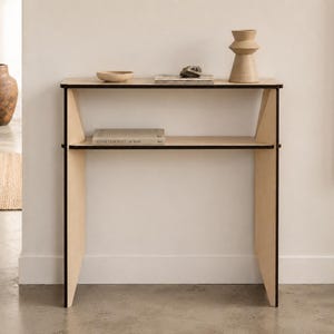 Könnte beinhalten: Ein heller Holzkonsolentisch mit zwei horizontalen Ablagen und dunkel abgesetzten Seitenstützen. Auf der oberen Ablage befinden sich eine kleine Schale, ein Stapel Bücher, ein Stein und eine Vase. Ein Buch liegt auf der unteren Ablage.
