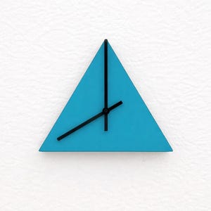 Puede incluir: Un reloj de pared moderno con una esfera triangular turquesa y manecillas negras. El reloj está montado sobre una superficie blanca texturizada. Las manecillas indican la hora.
