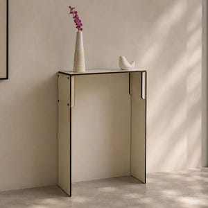 May include: A tall, narrow console table with a cream-coloured surface and dark edges. The table features a flat top and two vertical side panels. A tall, slender, two-toned vase containing purple flowers and a small white bird figurine sit on top.