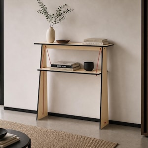 May include: A contemporary wooden console table with two shelves, black edging, and red supports. It features books, a vase with foliage, and a small bowl. The design is minimalist, set against a neutral wall, and is approximately 100cm in height.