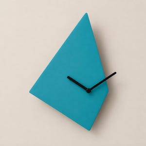 May include: A wall clock with an asymmetrical, geometric, teal-colored face. The clock features two simple, thin black hands positioned at the center of the angular shape, indicating the time without any numbers or markers on the clock face.