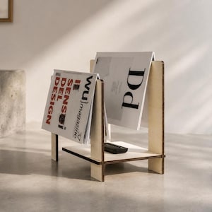 May include: A light-colored wooden magazine rack with a flat base and two vertical side panels. The rack holds two magazines, one featuring the text 'WU magazine' and 'SENSI DEL DESIGN', and the other displaying a large letter 'Dd' on the cover.
