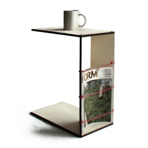 Pode incluir: Uma mesa auxiliar de madeira branca com tampo preto e um porta-revistas. O porta-revistas é de madeira e tem uma banda elástica vermelha que segura uma revista intitulada "FORM". Uma caneca de cerâmica branca está sentada no topo da mesa.
