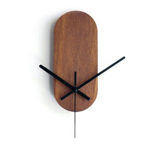 Könnte beinhalten: Eine minimalistische Wanduhr mit einem ovalen Holzgesicht und schwarzen Zeigern. Das Zifferblatt der Uhr hat eine warme braune Farbe, und die Zeiger sind dünn und elegant. Die Uhr ist etwa 30 cm hoch.