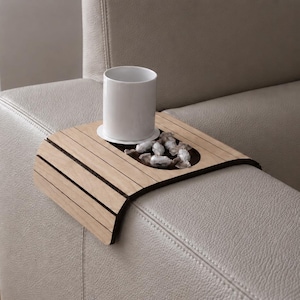 Puede incluir: Una bandeja de brazo de sofá de madera con un portavasos incorporado y un pequeño cuenco lleno de piedras decorativas. Una taza de cerámica blanca se encuentra en el portavasos. La bandeja es de color marrón claro y descansa sobre un brazo de sofá de cuero gris claro.