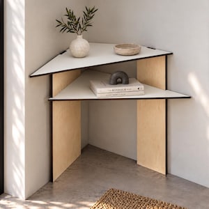 Op de afbeelding: Een hoekplank met twee niveaus, voorzien van verticale steunen van licht hout en witte driehoekige oppervlakken met donkere randen. De bovenste plank bevat een kleine witte vaas met groen en een kom, de onderste plank bevat twee boeken en een grijs boogobject.