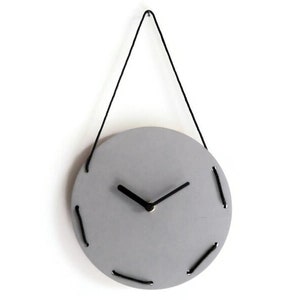 Könnte beinhalten: Eine minimalistische, runde Wanduhr in Hellgrau. Die Uhr hat schwarze Zeiger und wird von einer schwarzen Schnur gehalten. Das Zifferblatt ist mit schwarzen Nähten am Umfang verziert, was ein einzigartiges Designelement hinzufügt.