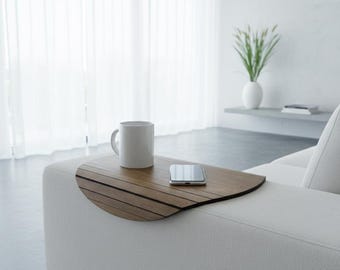 Può includere: Un vassoio flessibile in legno per braccioli con design a doghe appoggiato su un divano bianco. Il vassoio presenta una finitura con venature del legno marrone scuro e sostiene una tazza in ceramica bianca e uno smartphone rettangolare.