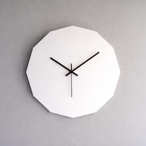 May include: A modern, white wall clock with a geometric, multi-sided face. The clock features black hands indicating the time against the white background. The clock is mounted on a gray wall.