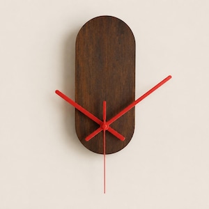 May include: A wall clock featuring a dark brown, vertically oriented oval wooden face. The clock has four bright red hands of varying lengths extending from the center, with one long red hand pointing straight down from the central mechanism.