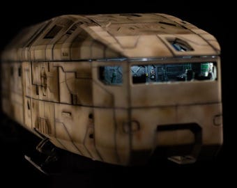 May include: A miniature sci-fi transport vehicle model featuring a weathered tan and brown camouflage paint scheme. The exterior has moulded panel lines and geometric details. The cockpit window reveals a detailed interior with illuminated controls.