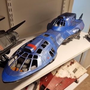 Op de afbeelding: Een blauw ruimteschipmodel met een gedetailleerd ontwerp, met een cockpit en meerdere ramen. Het model heeft rode en zwarte accenten en wordt tentoongesteld op een witte plank. Andere modellen zijn zichtbaar op de achtergrond.