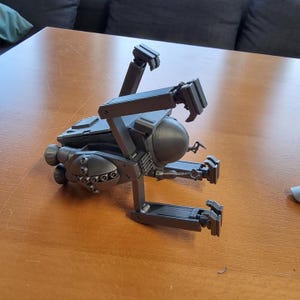 May include: A dark gray, mechanical model with articulated arms and a spherical component. The model has multiple sections with detailed features, including what appears to be a series of small circular lights. The object is resting on a wooden surface.