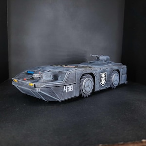 Puede incluir: Modelo gris oscuro de un vehículo futurista con un perfil bajo y seis ruedas. El vehículo tiene una torreta en la parte superior y el número "438" en el lateral. Un escudo es visible en el lateral. El vehículo está sobre un fondo oscuro.