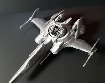 Puede incluir: Un modelo de nave espacial de plástico de color plateado con una nariz puntiaguda, cuatro alas extendidas con cañones montados y una cabina central abierta que contiene una pequeña figura de piloto. El vehículo tiene una superficie mecánica detallada.