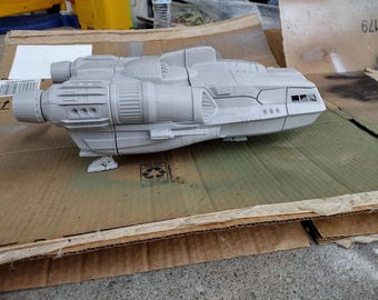 May include: A light grey, unpainted plastic model of a spaceship featuring a cockpit with visible windows, large side-mounted engine thrusters, and a segmented hull design. The spacecraft is shown from a side profile view, displaying intricate paneling.