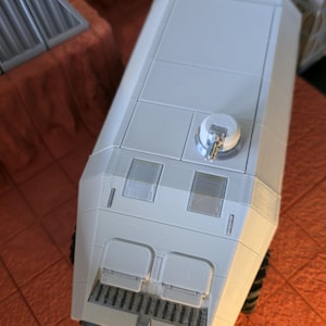 以下が含まれることがあります： 薄いグレーの角張った3Dプリント製の車両モデル。平らな屋根に小さな円形の砲塔、2つの長方形のフロントハッチ、フロントグリル、側面の車輪が特徴で、高い位置からのアングルで撮影されています。