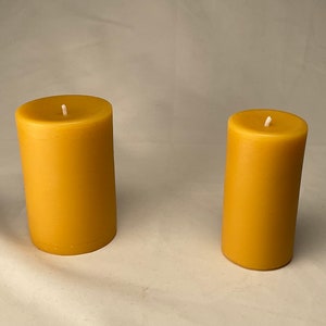 Peut inclure: Deux bougies en cire d'abeille jaune, l'une plus grande que l'autre, sur une surface blanche.