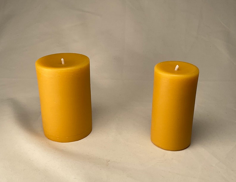 Peut inclure: Deux bougies en cire d'abeille jaune, l'une plus grande que l'autre, sur une surface blanche.