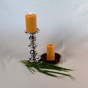 Peut inclure: Deux bougies, l'une sur un chandelier argent&eacute; et l'autre dans un plat brun, sont allum&eacute;es et br&ucirc;lent. Les bougies sont de couleur jaune p&acirc;le. Un bouquet d'herbe verte se trouve au premier plan.