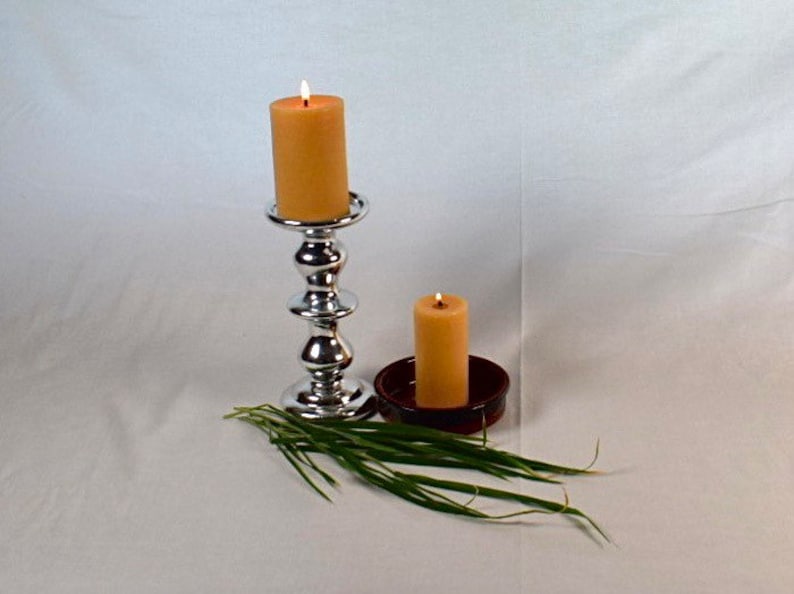 Peut inclure: Deux bougies, l'une sur un chandelier argent&eacute; et l'autre dans un plat brun, sont allum&eacute;es et br&ucirc;lent. Les bougies sont de couleur jaune p&acirc;le. Un bouquet d'herbe verte se trouve au premier plan.