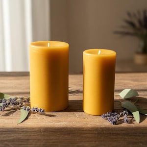 May include: Two yellow cylindrical candles of different sizes sit on a wooden surface. The candles are unscented and have a smooth texture. Lavender sprigs and green leaves are arranged around the candles, adding a natural touch.