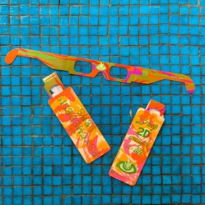 May include: A pair of psychedelic 3D glasses with a pink, orange, and gold design. The glasses are folded and have a small card attached that says "Psychedelic Lens" and "Bring 2D images to life!"