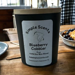 Pode incluir: Uma vela preta com tampa de madeira. O rótulo diz "Simple Scents Blueberry Cobbler" com um gráfico de cavalo. A vela tem 255 gramas e é feita de uma mistura de cera de soja 100%. O site 4ddsimplescents.com também está listado.