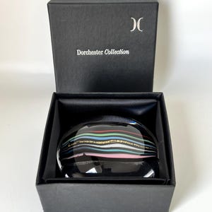 Puede incluir: Un pisapapeles negro con rayas horizontales rojas, azules, verdes, rosas y doradas. El pisapapeles está dentro de una caja negra con las palabras "Dorchester Collection" impresas en la tapa.