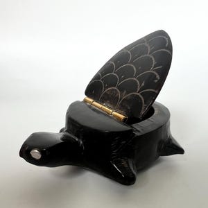 May include: A black turtle-shaped box with an open lid. The lid is hinged with gold-colored metal and features a carved scale pattern. The turtle's head and feet are visible, and the box is set against a white background.
