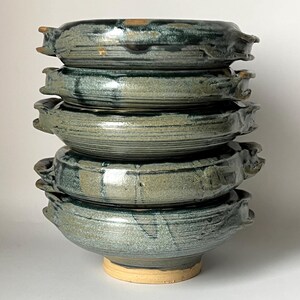 Può includere: Un set di cinque ciotole in ceramica con una glassa verde e una base marrone chiaro. Le ciotole hanno un aspetto unico e artigianale e sono perfette per servire una varietà di piatti.