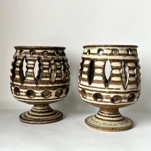 May include: Two ceramic candle holders with a brown and cream striped design. The holders have a pedestal base and a cylindrical body with cut-out shapes. The top rim has circular openings.