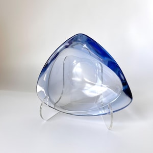 Op de afbeelding: Een driehoekige, heldere glazen kom met een diepblauwe rand. De kom wordt gepresenteerd op een heldere acryl standaard. Het ontwerp van de kom heeft een gelaagd effect, waardoor diepte en visuele interesse ontstaat. De kom is leeg.