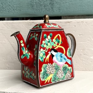 Puede incluir: Una pequeña tetera de cloisonné roja con un diseño de pájaro y flores. La tetera tiene una tapa con un pomo y un pico. La tetera está decorada con esmalte verde, amarillo, azul y blanco.