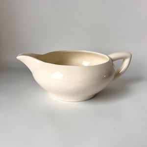 Op de afbeelding: Crèmekleurige keramische juskom met een afgeronde body, een schenktuit en een handvat. De juskom heeft een glanzende afwerking en een licht gebarsten uiterlijk, wat duidt op leeftijd of een vintage esthetiek. Het ontwerp is eenvoudig en klassiek.