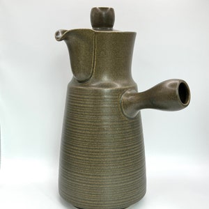 Può includere: Una caffettiera in ceramica marrone con una superficie testurizzata e un coperchio. La caffettiera ha un manico e un beccuccio.