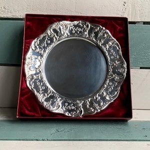 Puede incluir: Un plato de servicio de plata con un diseño ornamentado y en relieve. El plato está en una caja de terciopelo rojo.