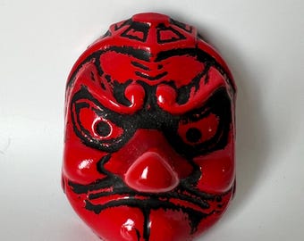 May include: A red and black Japanese Noh mask with a fierce expression. The mask has bold black lines outlining the eyes, eyebrows, and mouth, creating a striking contrast against the red base. The mask is a traditional art form.