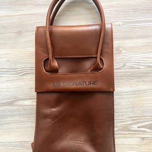 May include: A brown leather wine tote bag with two handles. The bag has a rectangular shape with a flap closure and the text "LG SIGNATURE" printed on the front.