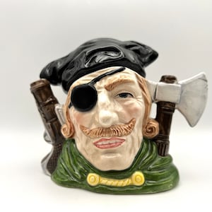 May include: A ceramic figurine of a man with a black eye patch, black hat, and green jacket. He is holding an axe and a gun in his hands.