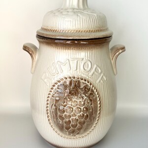 Könnte beinhalten: Eine weiße Keramik-Dose mit braunem Deckel und einem braunen Traubendesign. Die Dose hat das Wort "Rumtopf" auf der Seite eingeprägt.