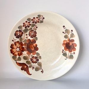 Könnte beinhalten: Cremefarbene Keramikplatte mit floralem Design. Die Blumen sind in Braun- und Orangetönen mit grünen Blättern und Stielen bemalt. Die Platte hat ein leicht rissiges Aussehen.