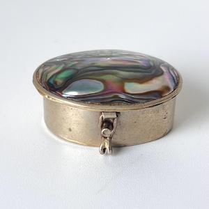 May include: A round metal pill box featuring an iridescent abalone shell inlay on the lid. The container has a silver-toned finish and a small hinged latch mechanism on the front side to secure the lid, showcasing swirling green and purple hues.