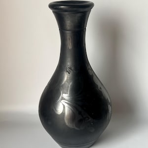 Op de afbeelding: Een zwarte keramische vaas met een smalle hals en een brede, ronde basis. De vaas is versierd met een zilveren bloemenontwerp.