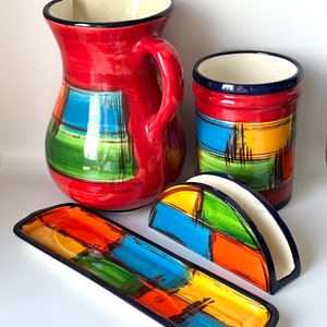 May include: A set of colorful ceramic kitchenware featuring a pitcher, a napkin holder, a utensil holder, and a rectangular serving dish. The pieces are decorated with a geometric pattern of red, orange, yellow, green, and blue.