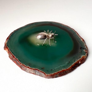 Puede incluir: Una figura de insecto de metal en tono plateado pulido con patas delgadas y un cuerpo liso y redondeado, colocada sobre una rodaja circular y plana de piedra ágata verde con un borde exterior marrón oscuro y rojizo y un centro más claro.