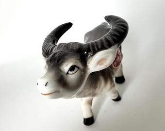 May include: A small, ceramic figurine of a gray and white water buffalo with black horns. The figurine has a red and white label on its side that reads "Bone China".